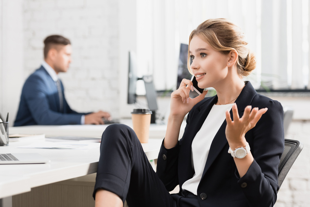 Positive businesswoman gesturing, while talking on mobile phone at workplace on blurred background - Photo, Image