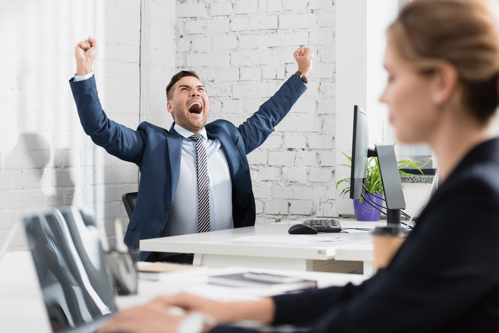 Excited executive with yes gesture sitting at workplace with blurred colleague on foreground - Photo, Image