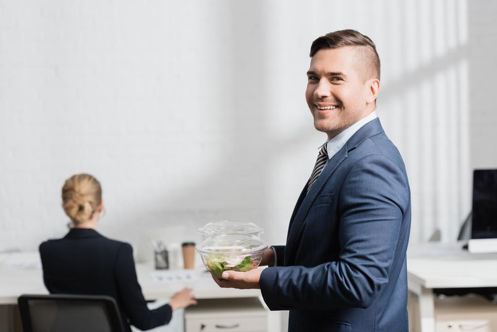 Hombre de negocios sonriente con comida en tazones de plástico mirando a la cámara, mientras está de pie en la oficina sobre un fondo borroso - Foto, imagen
