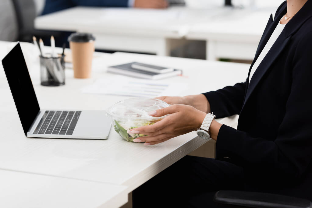 Vista cortada de empresária segurando tigela de plástico com refeição, enquanto sentado no local de trabalho em fundo turvo - Foto, Imagem