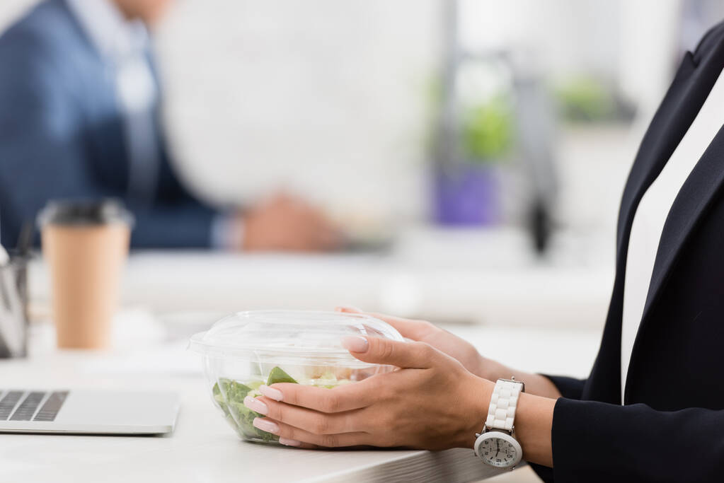 Vista recortada de una mujer ejecutiva sosteniendo un tazón de plástico con comida en el lugar de trabajo sobre un fondo borroso - Foto, imagen