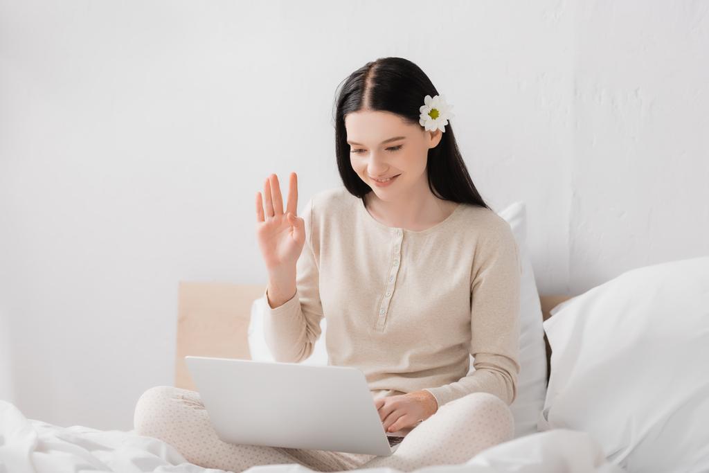 femme heureuse avec vitiligo et fleur dans les cheveux agitant la main tout en ayant appel vidéo sur ordinateur portable dans la chambre  - Photo, image