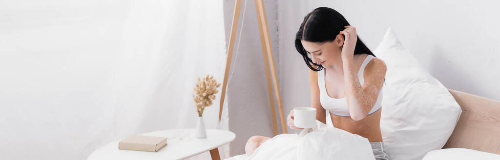 улыбающаяся женщина с витилиго держа чашку кофе во время фиксации волос в постели, баннер - Фото, изображение