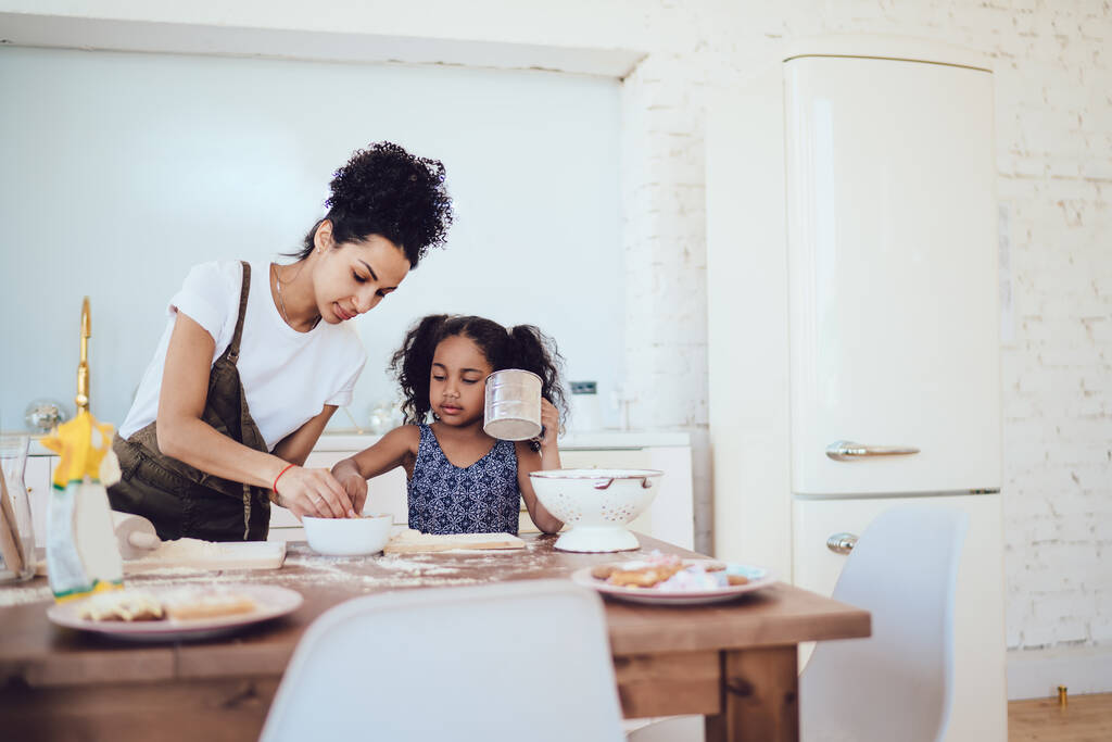 Joven madre rizada afroamericana en traje causal de pie con la niña en la mesa y cocinar en la cocina en casa  - Foto, imagen