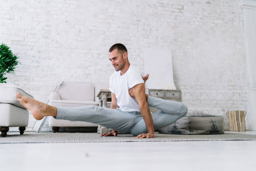 Smiling hipster guy in casual wear sitting in asana stretching muscles on morning training enjoying healthy lifestyle, handsome positive man recreating and training staying at home in apartment - Photo, Image