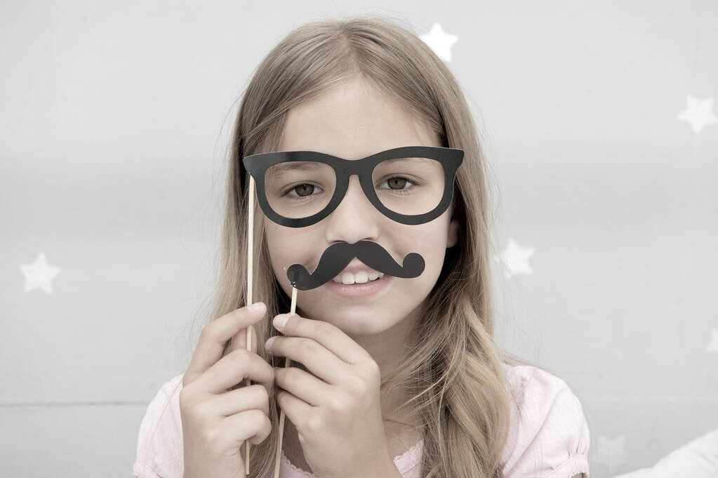 Девушка позирует с фотобудкой реквизит очки и усы в спальне. Девочка в очках. Ребенок подражает мужественности или хочет выглядеть как ее отец. Парень с усами и очками - Фото, изображение
