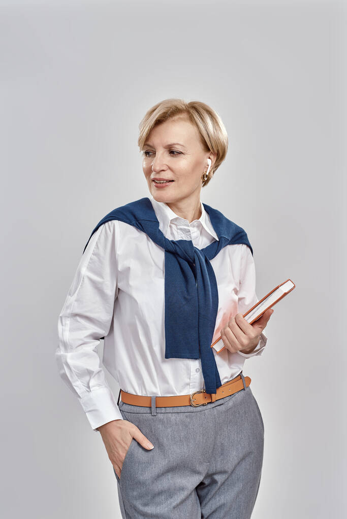 Portrait d'une élégante femme caucasienne d'âge moyen portant des vêtements d'affaires regardant de côté, tenant son carnet tout en se tenant isolée sur fond gris - Photo, image