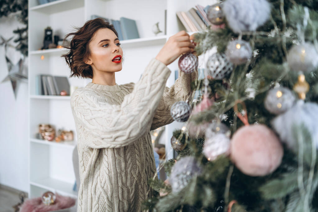 Natal, Ano Novo. Morena bonita e feliz em suéter quente decorando árvore de Natal em casa - Foto, Imagem