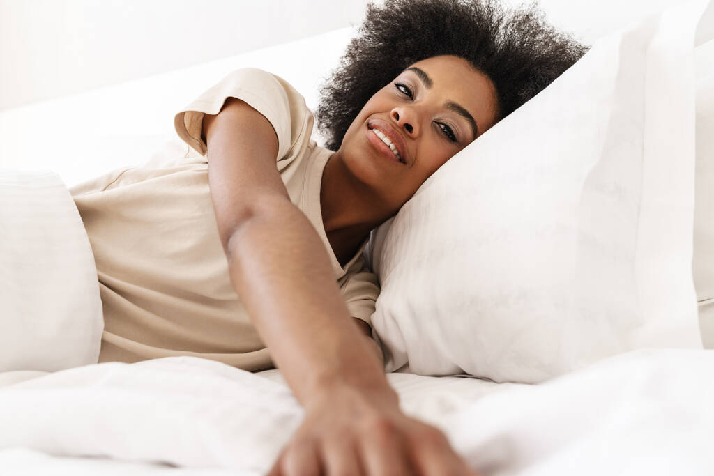 Εικόνα του νεαρού ωραίο αφροαμερικανό κορίτσι χαμογελά στην κάμερα μετά τον ύπνο σε λευκό κακό - Φωτογραφία, εικόνα