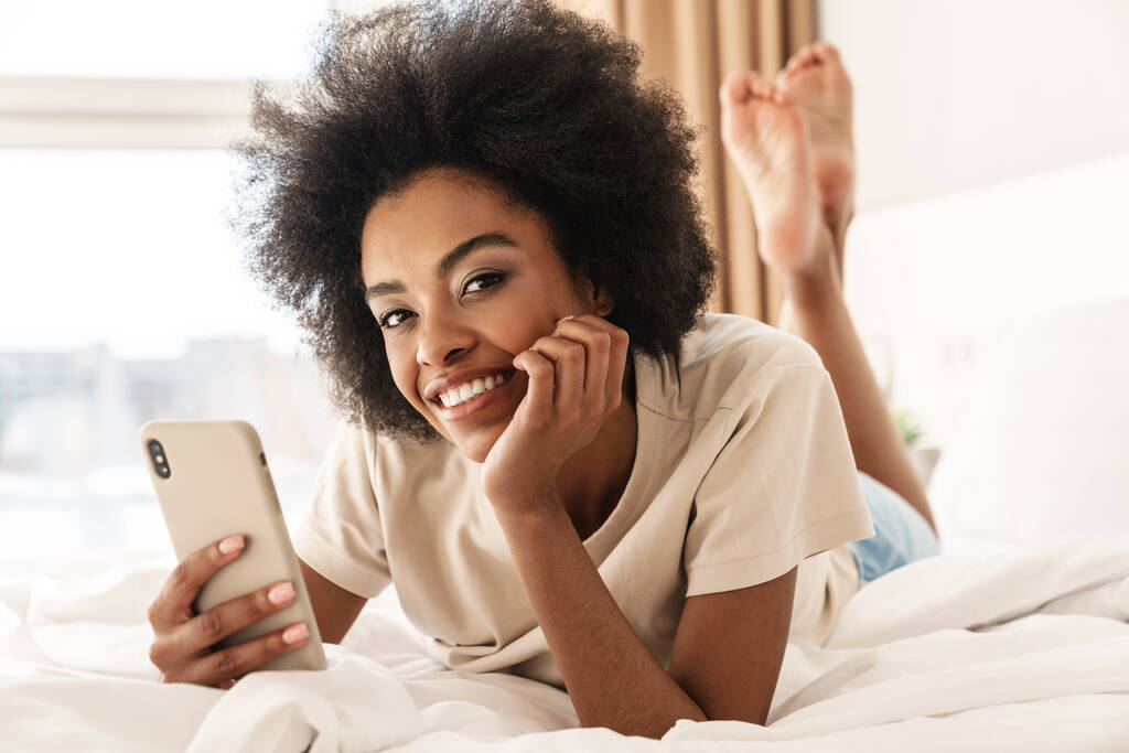 Jonge glimlachende Afrikaanse vrouw die sms 'jes stuurt, in de slaapkamer ligt - Foto, afbeelding