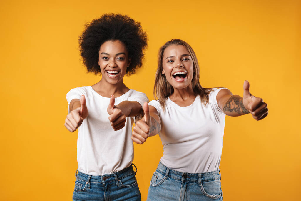 Зображення позитивних молодих багаторасових дівчат друзів, які позують ізольовані на жовтому тлі стіни, показуючи великі пальці вгору жестом
 - Фото, зображення