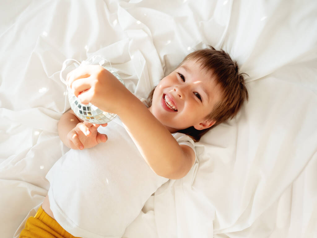 Kleiner Junge spielt mit glitzernder Spiegel-Discokugel im Bett und lacht glücklich. Fröhliches Kleinkind. Verspieltes Kind und sonniger Morgen im gemütlichen Zuhause. - Foto, Bild