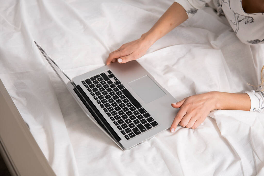Draufsicht einer Frau, die mit Laptop auf einem Bett sitzt. Arbeiten zu Hause Konzept. Mädchen arbeitet an ihrem Laptop im Bett mit weißer Bettwäsche. Entspannter Arbeitsprozess. Zu Hause bleiben. - Foto, Bild