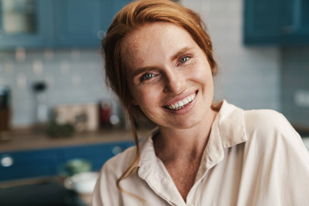 Obrázek šťastné zrzky žena usmívá a dívá se na kameru, zatímco stojí doma kuchyně - Fotografie, Obrázek