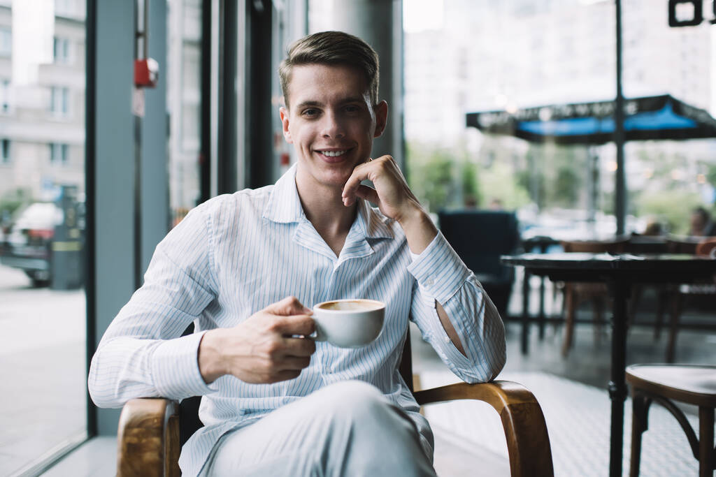 Positive successful man sitting at wooden table in comfort armchair while drinking hot beverage indoors at modern cafeteria at daytime - Photo, Image