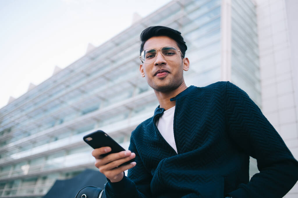 Πορτρέτο του θετικού hiddy hipster τύπος σε μοντέρνα γυαλιά χρησιμοποιώντας το κινητό τηλέφωνο για την επικοινωνία σε απευθείας σύνδεση, όμορφος άντρας ντυμένος με κομψό ντύσιμο κοιτάζοντας κάμερα ξοδεύοντας χρόνο στην πόλη δρόμο - Φωτογραφία, εικόνα
