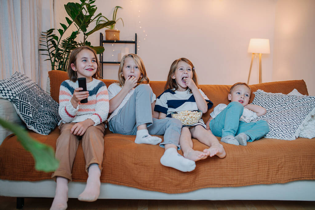 Παιδιά από 5 έως 10 ετών κάθονται στον καναπέ, βλέπουν τηλεόραση, τρώνε ποπ κορν - Φωτογραφία, εικόνα