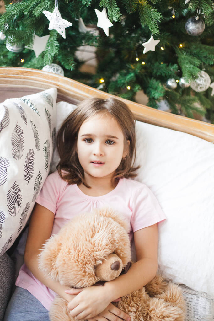 de kamer is ingericht voor het nieuwe jaar en Kerstmis, baby meisje op de bank knuffelen van een beer, jeugd, cadeau van ouders - Foto, afbeelding