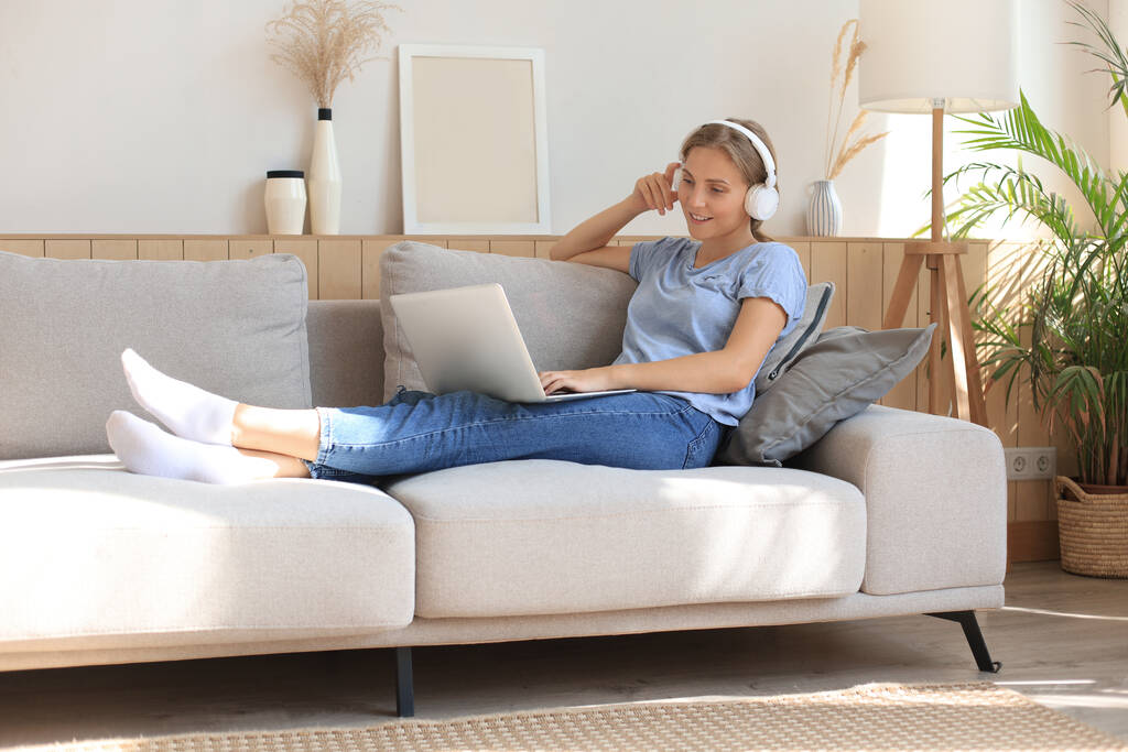 Улыбающаяся молодая женщина с наушниками и ноутбуком на диване - Фото, изображение