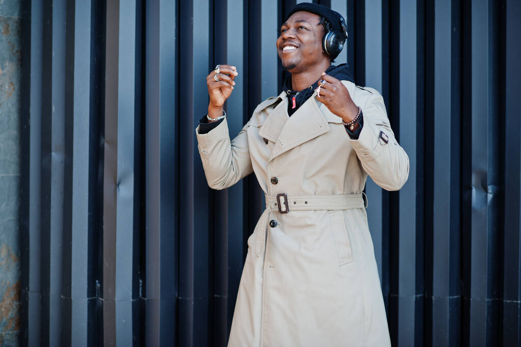 Αφροαμερικάνος με ακουστικά, φοράει καπέλο και παλτό ακούγοντας μουσική σε φόντο χάλυβα. - Φωτογραφία, εικόνα