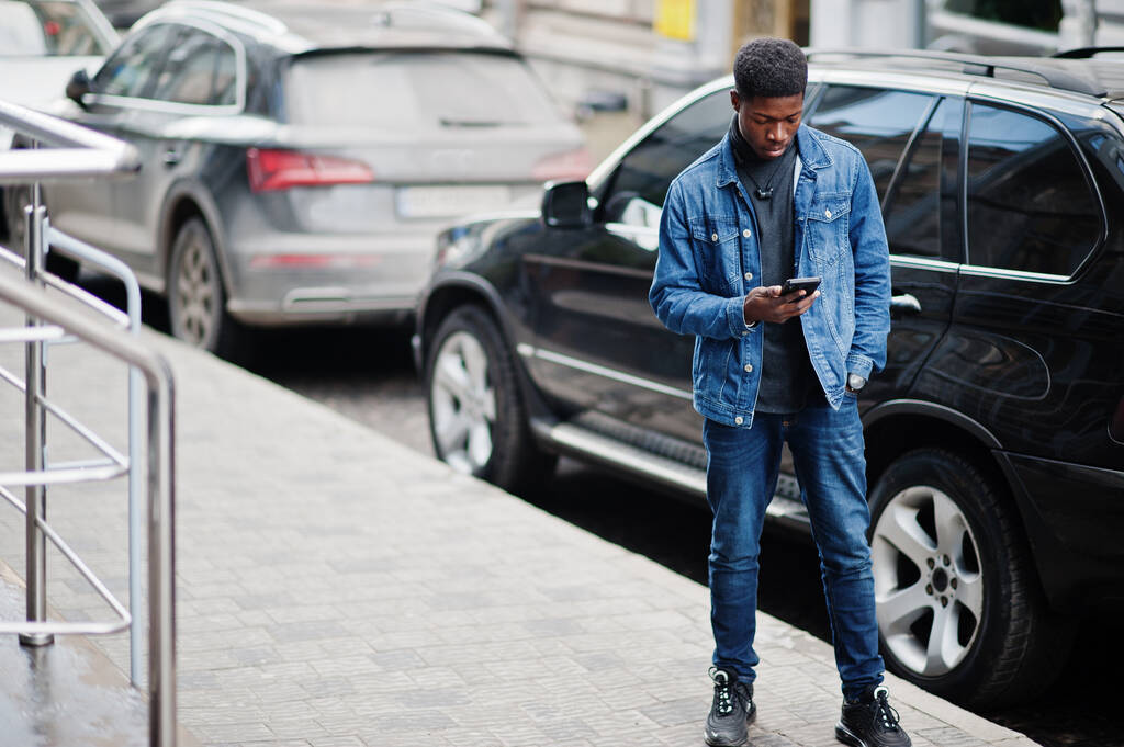 Afrikaner in Jeansjacke posierte draußen vor schwarzem Auto mit Handy in der Hand. - Foto, Bild