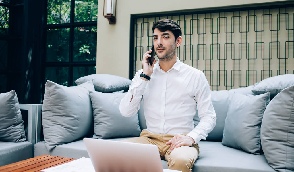 Από κάτω αποφασισμένος καλοντυμένος επιχειρηματίας επικοινωνεί στο κινητό τηλέφωνο, ενώ κάθεται στον καναπέ στο καφέ και κοιτάζοντας την κάμερα - Φωτογραφία, εικόνα