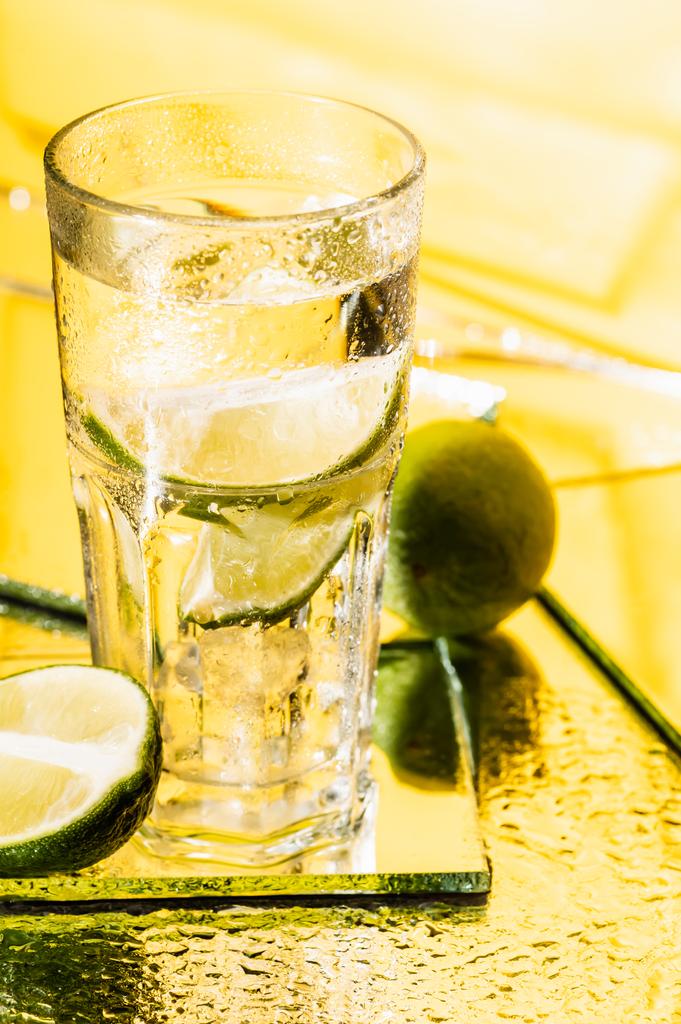 ποτήρι αλκοόλ κοκτέιλ κοντά σε φέτες ασβέστη σε κίτρινο  - Φωτογραφία, εικόνα