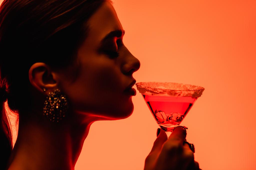 zijaanzicht van jonge vrouw met gesloten ogen drinken margarita cocktail op oranje  - Foto, afbeelding