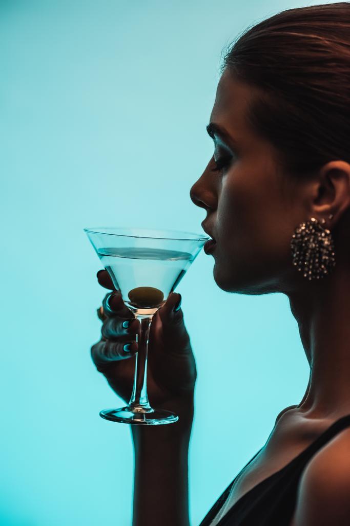 vue latérale de la femme tenant le verre et buvant martini isolé sur bleu - Photo, image