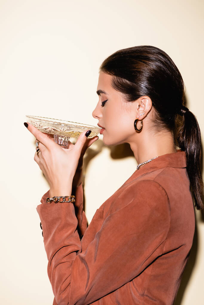 πλαϊνή όψη της μελαχρινής γυναίκας σε καφέ σακάκι πίνοντας σαμπάνια από γυαλί σε λευκό - Φωτογραφία, εικόνα