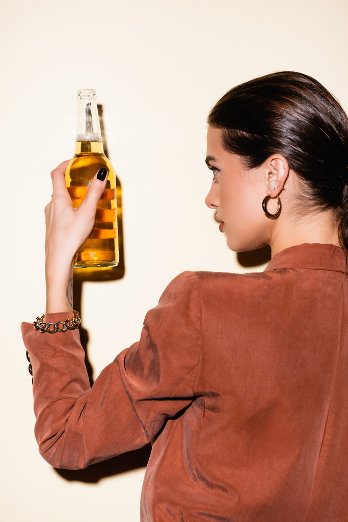 brünette Frau im braunen Blazer blickt auf Flasche mit Bier auf weißem Grund - Foto, Bild