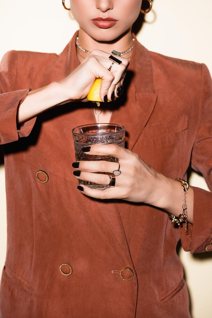 bijgesneden beeld van jonge vrouw in bruine blazer knijpen citroen in glas met alcohol cocktail op wit  - Foto, afbeelding