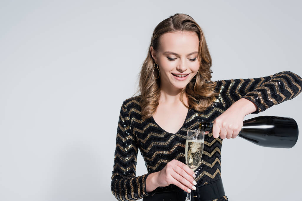 Ευτυχισμένη νεαρή γυναίκα κρατώντας το ποτήρι, ενώ ρίχνει σαμπάνια από το μπουκάλι απομονώνονται σε γκρι - Φωτογραφία, εικόνα