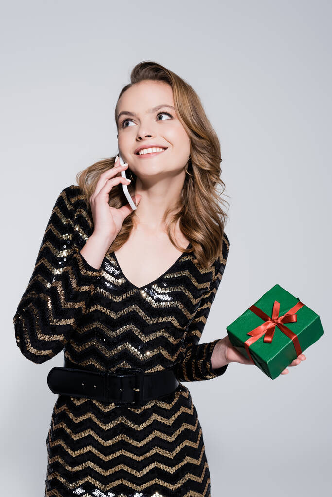 улыбающаяся молодая женщина, держащая в руках подарок на Рождество и разговаривая на смартфоне, изолированном от серого  - Фото, изображение