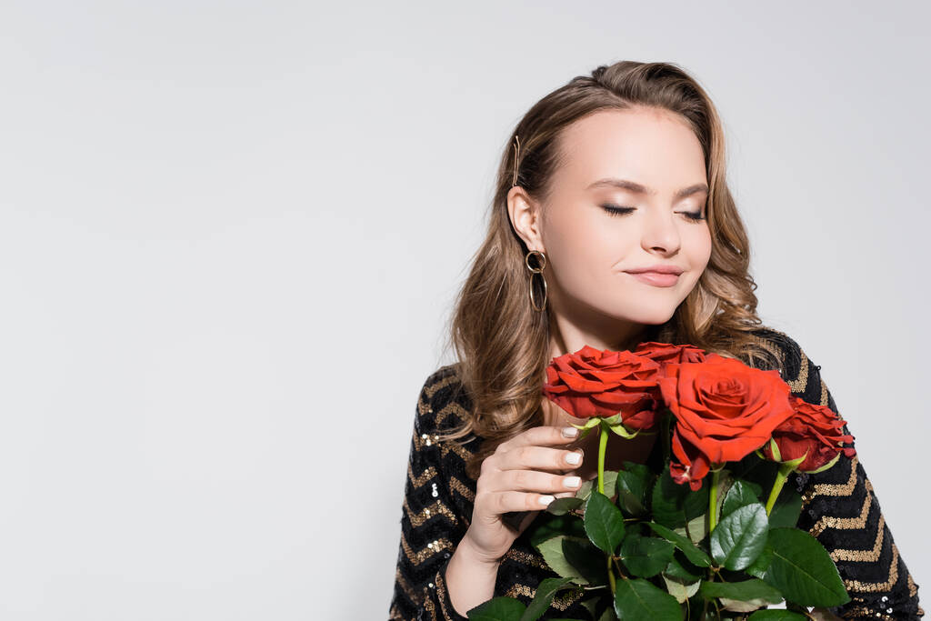 feliz joven mujer oliendo ramo de rosas rojas aisladas en gris - Foto, Imagen