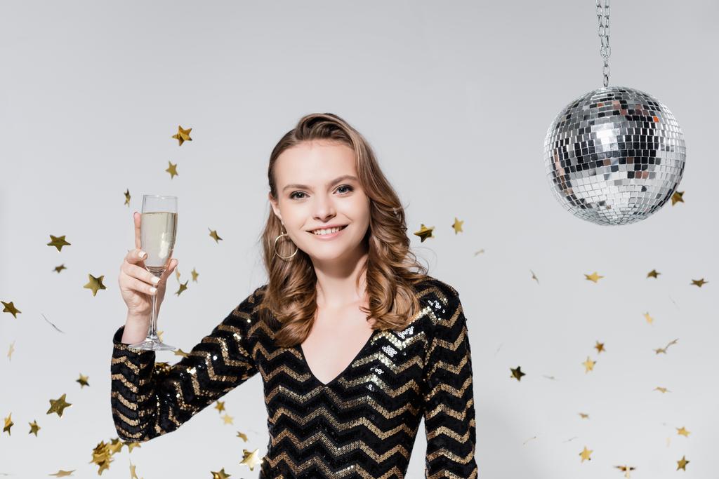 fröhliche junge Frau hält Glas Champagner in der Nähe von Discokugel und fällt Konfetti auf Grau - Foto, Bild