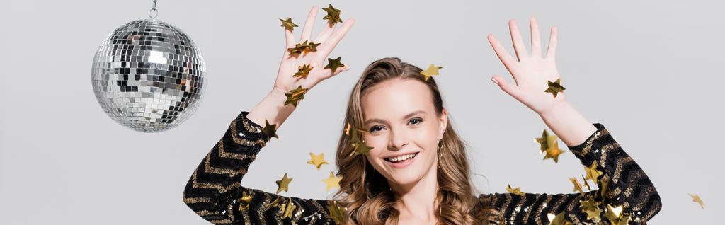 cheerful young woman standing near disco ball and falling confetti on grey, banner - Photo, Image