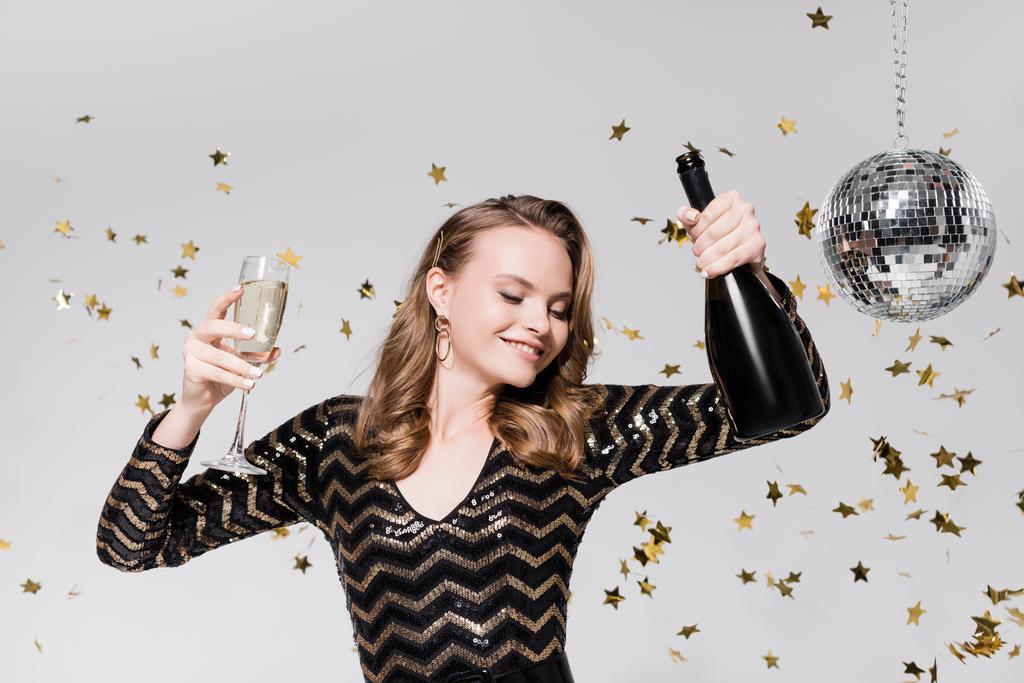 veselá mladá žena drží sklo a láhev šampaňského v blízkosti diskotéky míč a padající konfety na šedé - Fotografie, Obrázek