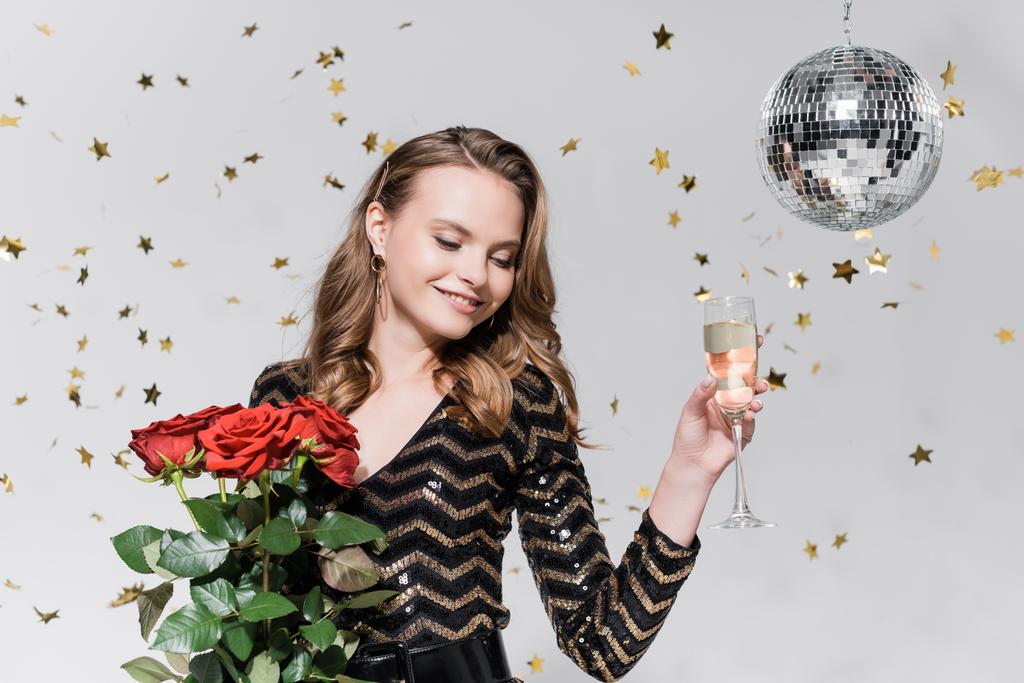 vrolijke jonge vrouw met rode rozen en glas champagne in de buurt van discobal en vallende confetti op grijs - Foto, afbeelding