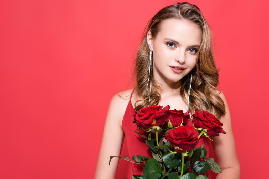 Χαρούμενη νεαρή γυναίκα που κρατάει τριαντάφυλλα και κοιτάζει την κάμερα στο κόκκινο  - Φωτογραφία, εικόνα