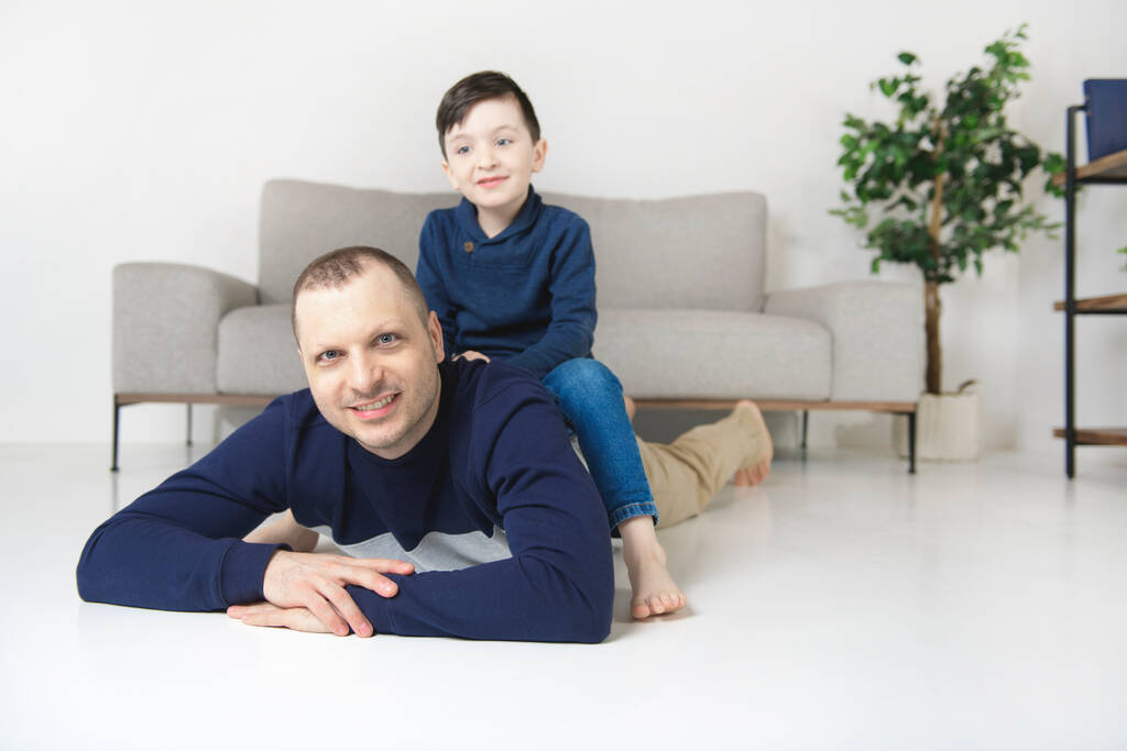 Χαριτωμένο αγόρι και ο όμορφος πατέρας της κοιτάζοντας κάμερα και χαμογελώντας ενώ στο σπίτι - Φωτογραφία, εικόνα