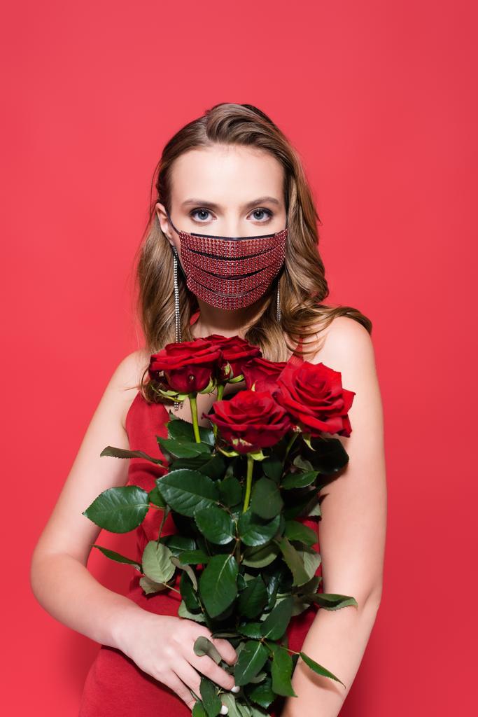 woman in protective mask with rhinestones holding roses and looking at camera on red  - Photo, Image