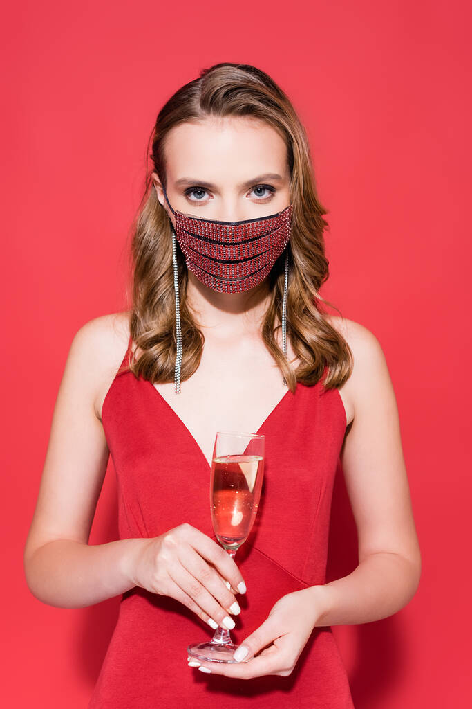γυναίκα με προστατευτική μάσκα με στρας που κρατά ένα ποτήρι σαμπάνια στο κόκκινο  - Φωτογραφία, εικόνα