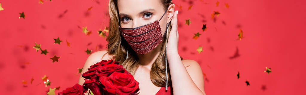woman in protective mask with rhinestones fixing hair near roses and confetti on red, banner - Photo, Image