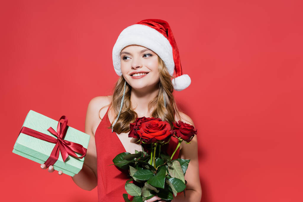 gelukkig jong vrouw in santa hoed houden rozen en gift box op rood - Foto, afbeelding