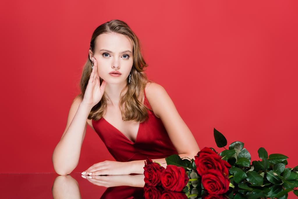 νεαρή γυναίκα κοιτάζει κάμερα κοντά σε τριαντάφυλλα σε κόκκινο - Φωτογραφία, εικόνα