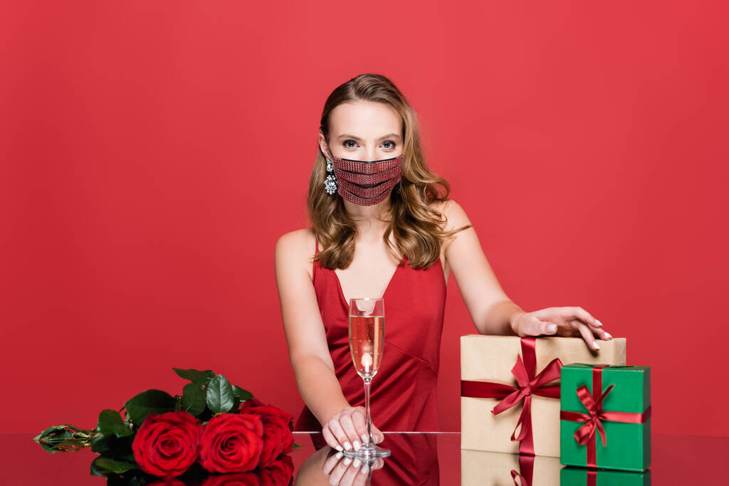 žena v ochranné masce s broušenými kameny se sklenicí šampaňského v blízkosti růží a vánočních dárků na červené - Fotografie, Obrázek