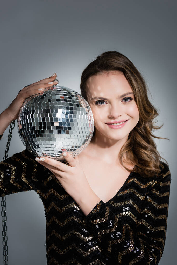 cheerful young woman looking at camera while holding disco ball on grey - Photo, Image