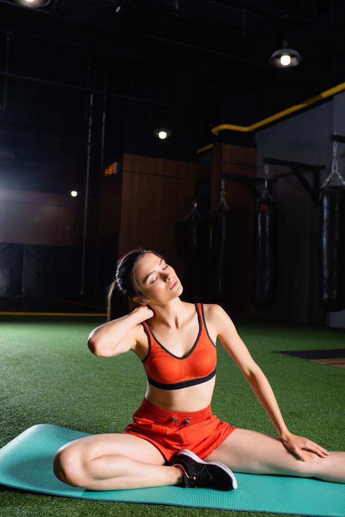 young sportswoman warming up neck while sitting on fitness mat with closed eyes - Photo, Image