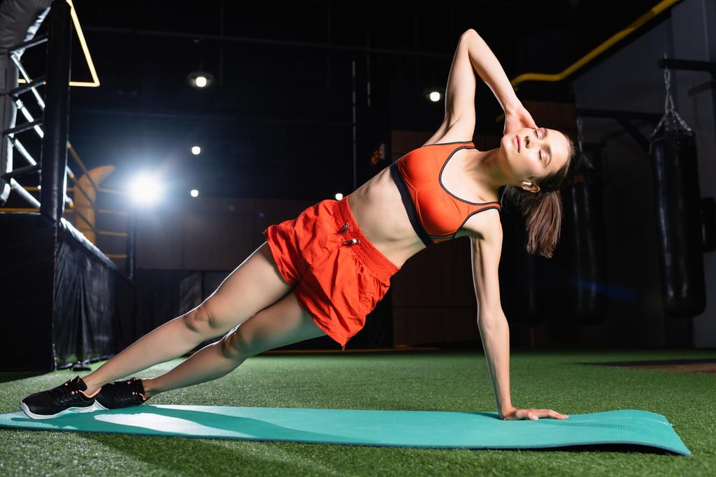 Sporcu kadın üstte ve şort spor salonunda kapalı gözlerle yan tahta egzersizi yapıyor. - Fotoğraf, Görsel